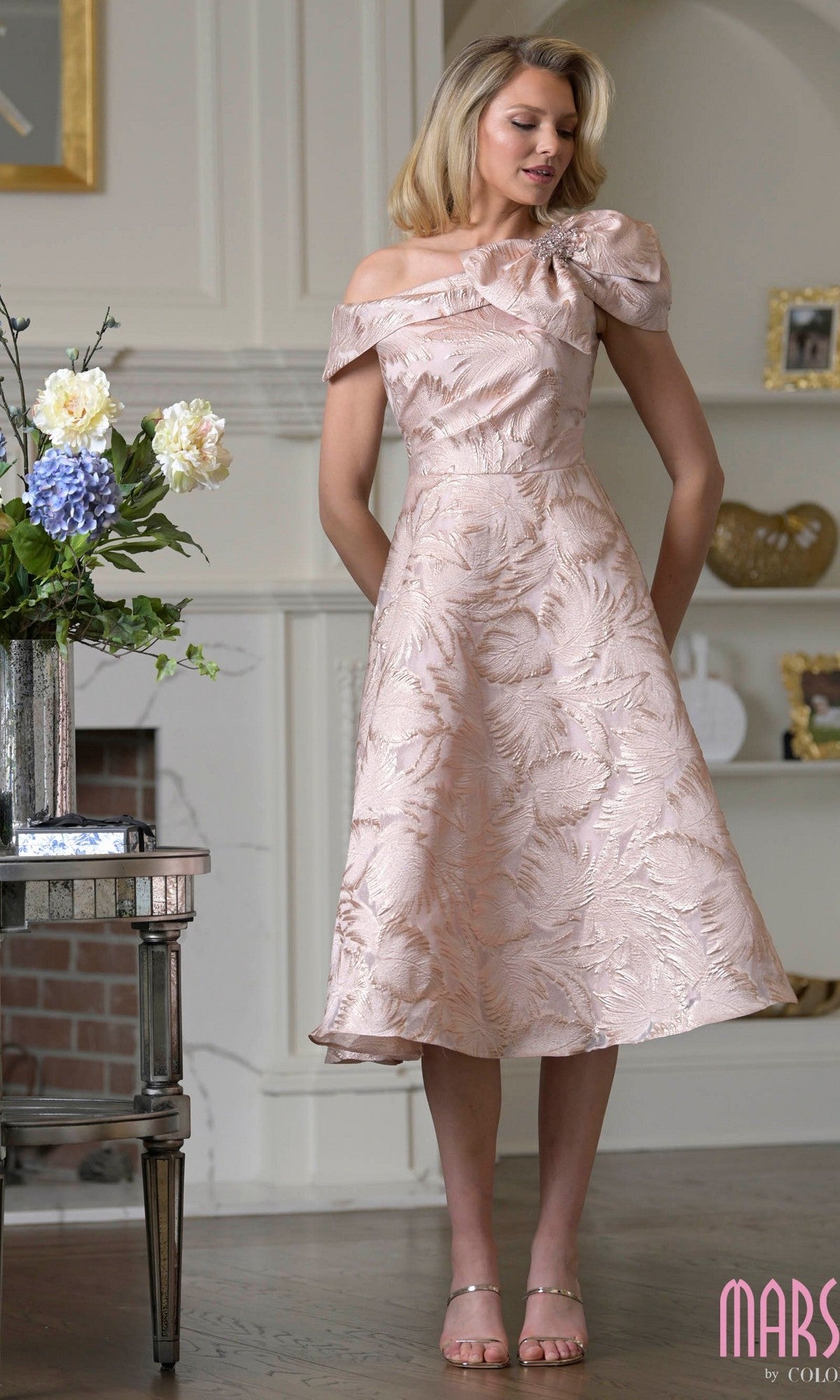 A woman stands indoors by a table with a vase of flowers, wearing the blush-colored knee length semi-formal dress Marsoni MV1413—an off-the-shoulder jacquard design with a textured leaf pattern and bow detail.